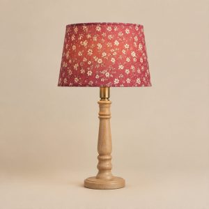 A table lamp with a pink floral shade lit above a turned light wood base, on a plain cream background