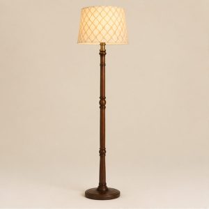 A floor lamp with a cream patterned shade lit on a turned dark wood column, on a plain cream background